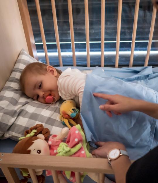 Nos professionnelles de la petite enfance accompagnent les enfants accueillis sur les temps de siestes. enfant dans son lit à barreau, avec une tétine, et une professionnelle qui pose la main sur lui